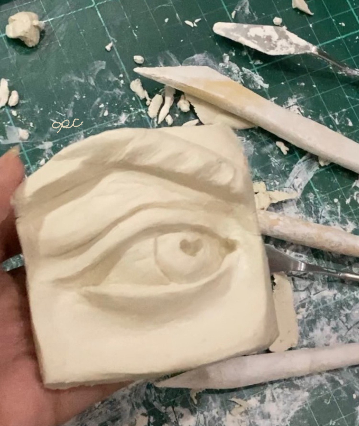 Pale clay relief of an eye surrounded by sculpting tools and clay scraps.
