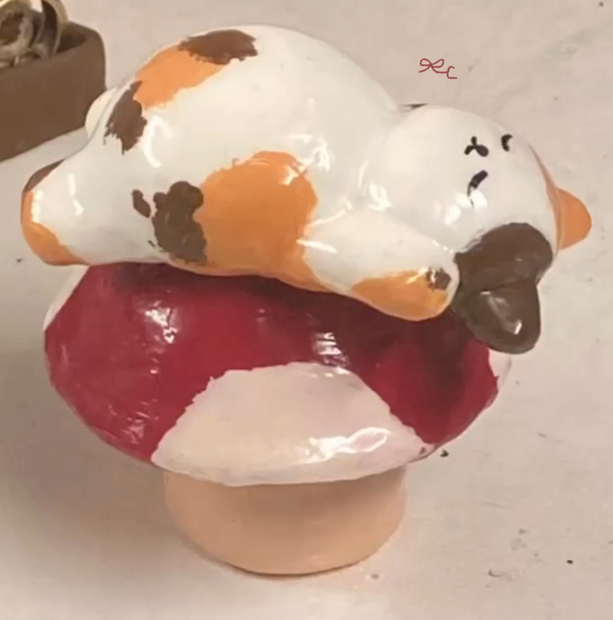 Painted clay cat figure resting on a red-and-white rounded base.
