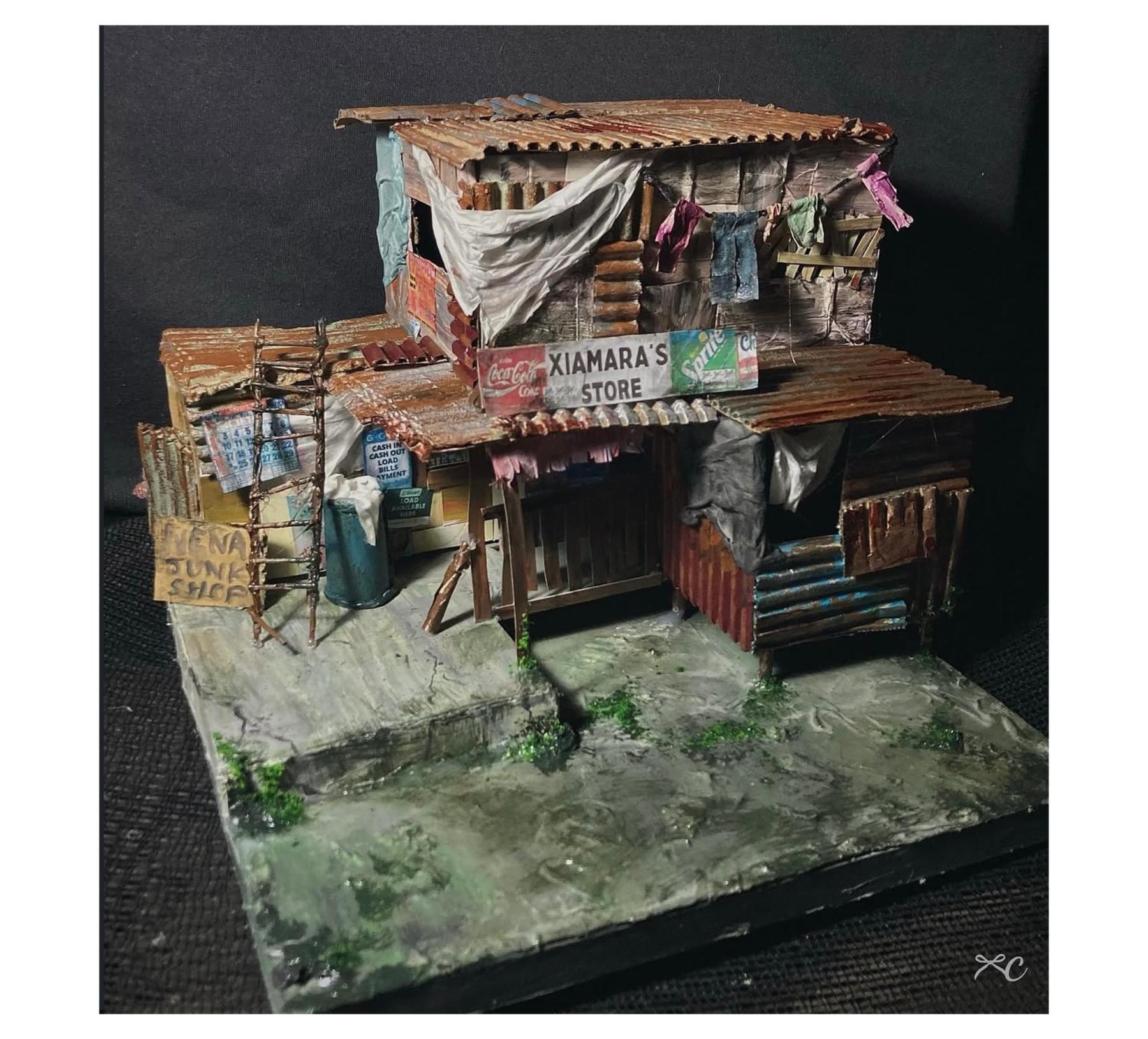 Diorama of makeshift homes with corrugated walls, grass, debris, and small figures.